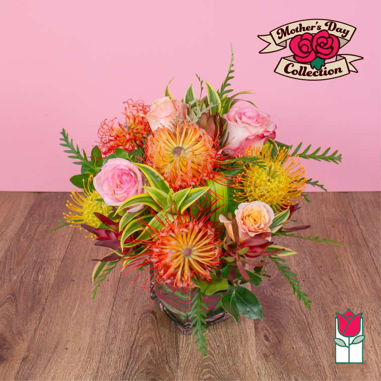Arrangement of orange and yellow protea with pink roses in a square glass vase on a wooden surface