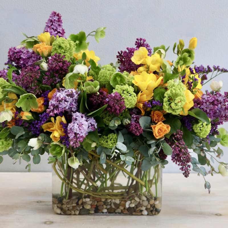 Lush mixed arrangement of purple, yellow, green, and orange flowers in a rectangular glass vase with pebbles.