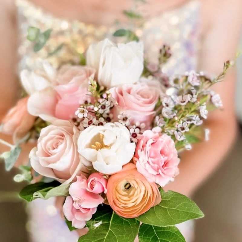 Person holding a small pastel bouquet of pink roses, white blooms, and peach ranunculus.