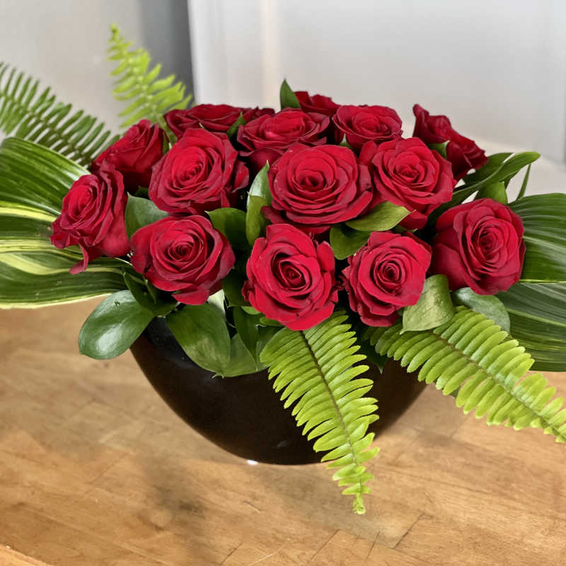 Red roses arranged in a black bowl with broad green leaves and ferns