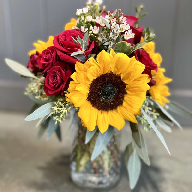 Arrangement of yellow sunflower, red roses, and small white blooms in a clear glass jar vase