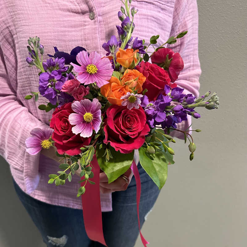 Handheld bouquet of bright pink roses, orange blooms, and purple flowers with a hot pink ribbon.