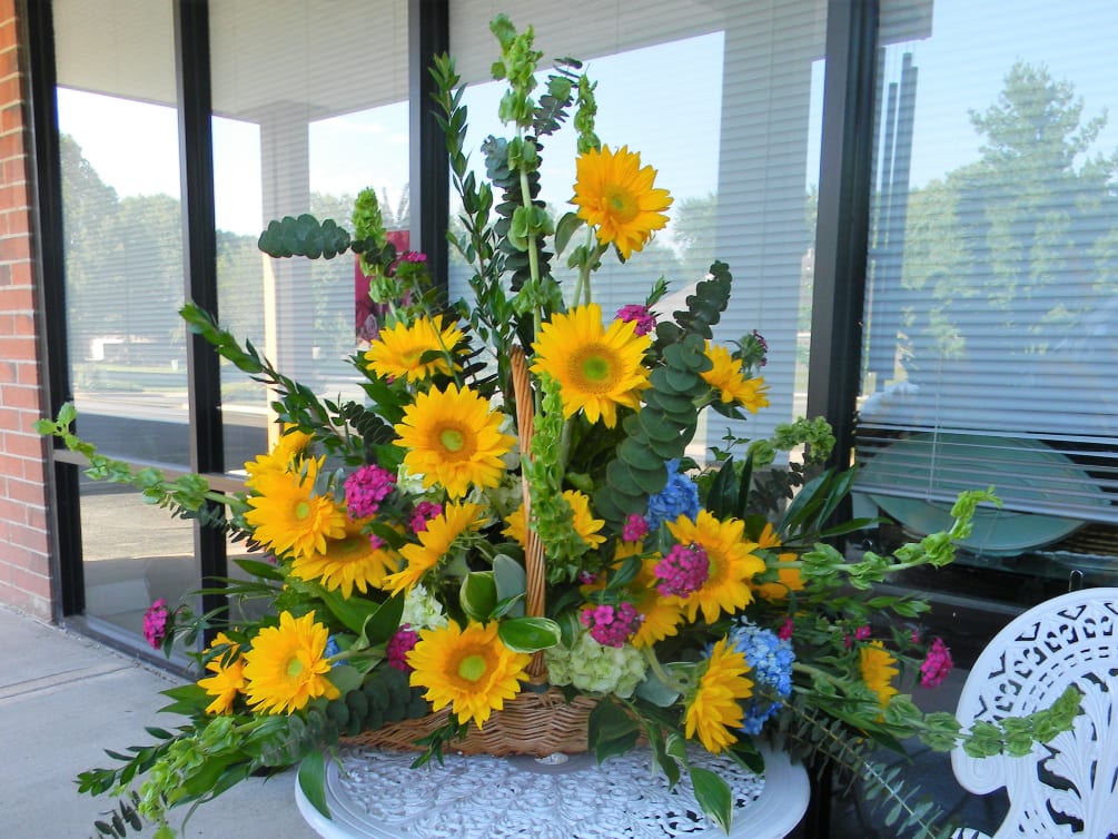 This fireside basket of sunflowers, orchid colored stock and seasonal foliage makes