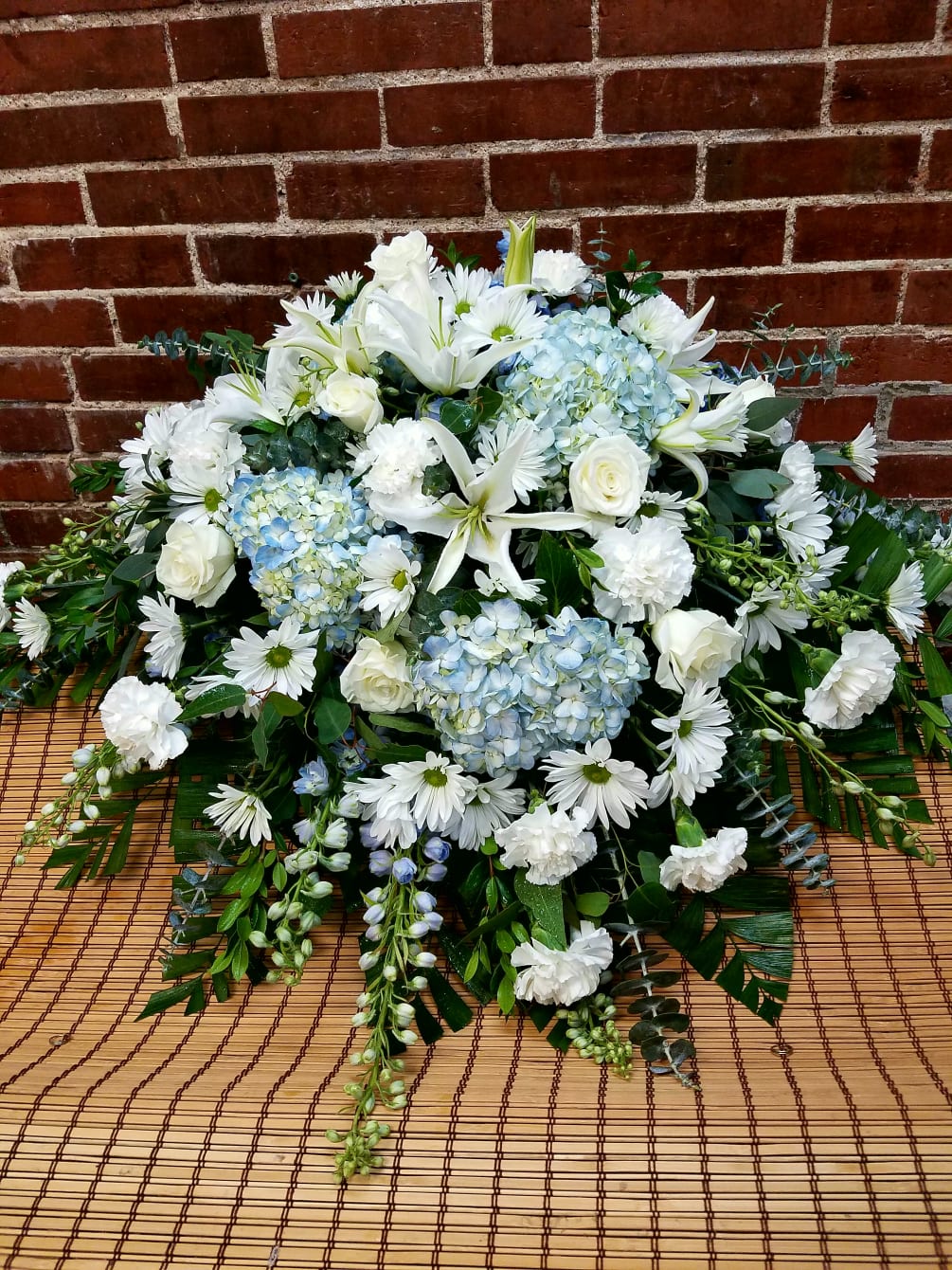 This casket adornment features blue hydrangeas, white lilies, blue delphinium, white roses