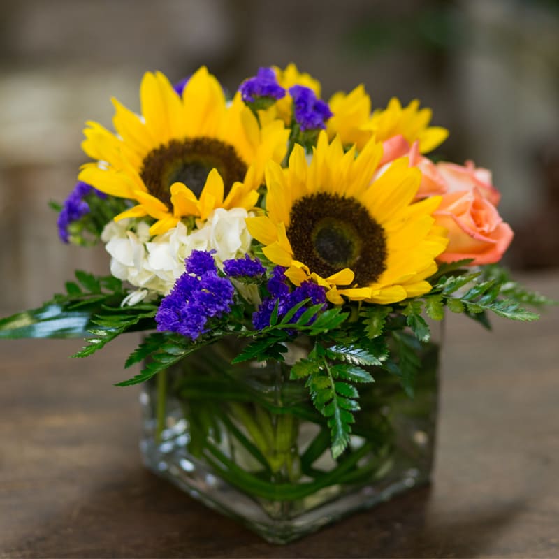 Clear square cube with vibrant sunflowers, white hydrangea, orange roses and purple