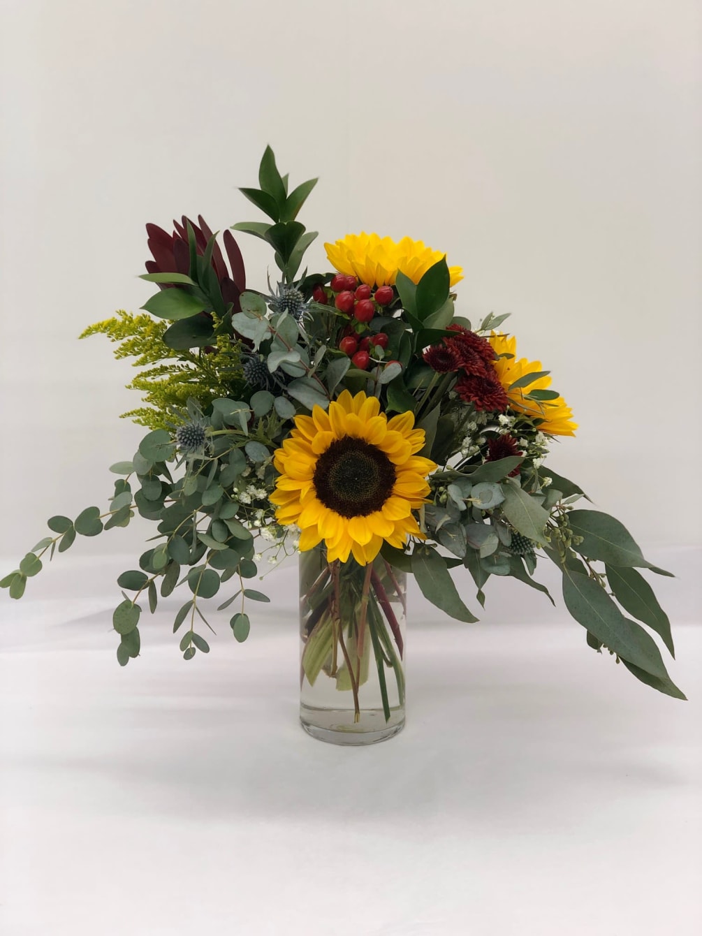 Organic arrangement of mainly assorted greenery with a touch of sunflowers, mums