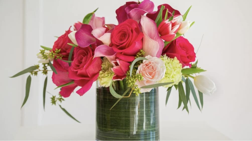 Pink callas arranged with roses and fillers in clear cylinder vase. Greens