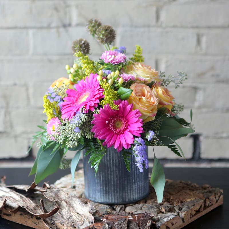 Gerberas, roses, solidago, and statice arranged in a tin container.