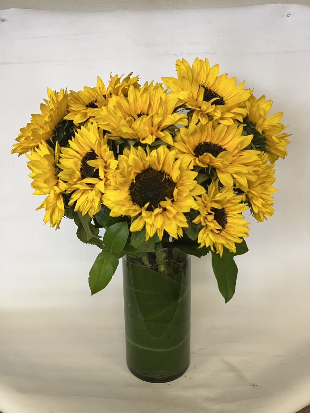 TALL VASE OF SUNFLOWERS WRAPPED IN GREENERY. 