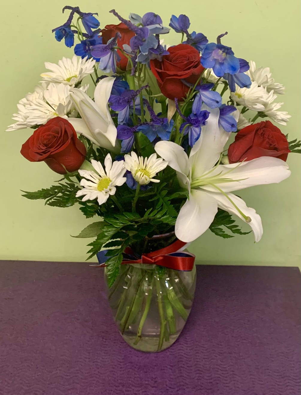 This arrangement includes white Hybrid Liles, red Roses, blue Delphinium, and white