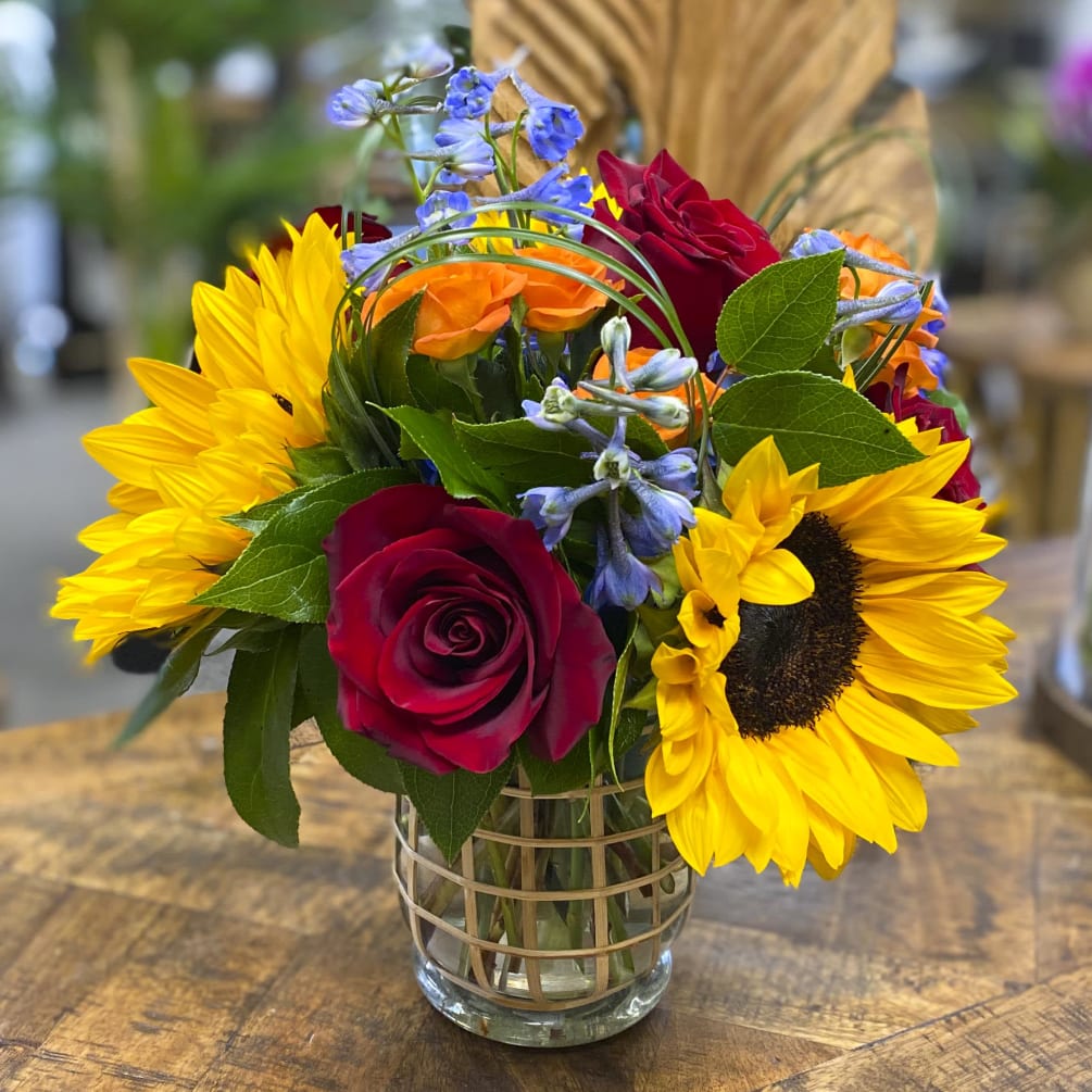 This beautiful arrangement is filled with sunflowers, roses, garden roses, blue delphiniums