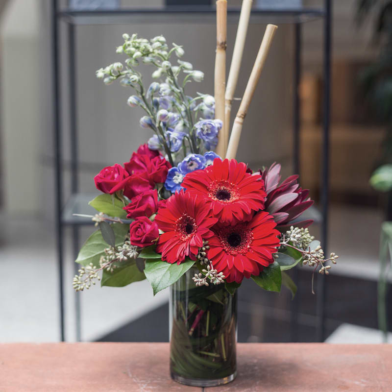 A artful display of Gerbera Daisies with Delphinium, Roses, exotic foliage&#039;s, and