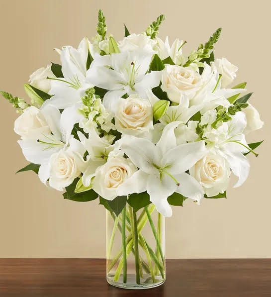 Round arrangement with white roses, lilies and snapdragons; with greenery. Presented in