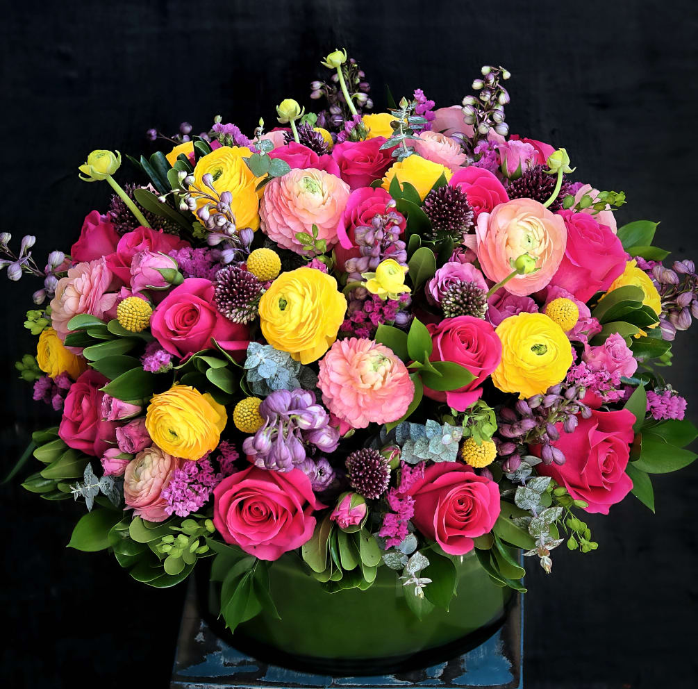 Bright centerpiece contains mixed florals such as roses, ranunculus, delphinium and others