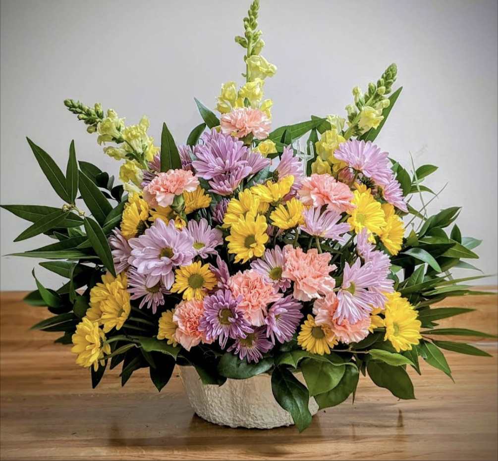 Spring inspired basket spray designed with daisies, carnations, and snapdragons.