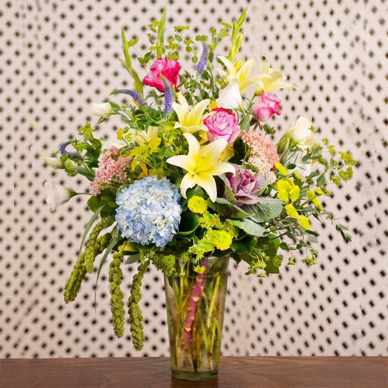 Blue hydrangeas, roses, lilies, mums, and delphinium in a tall glass vase.