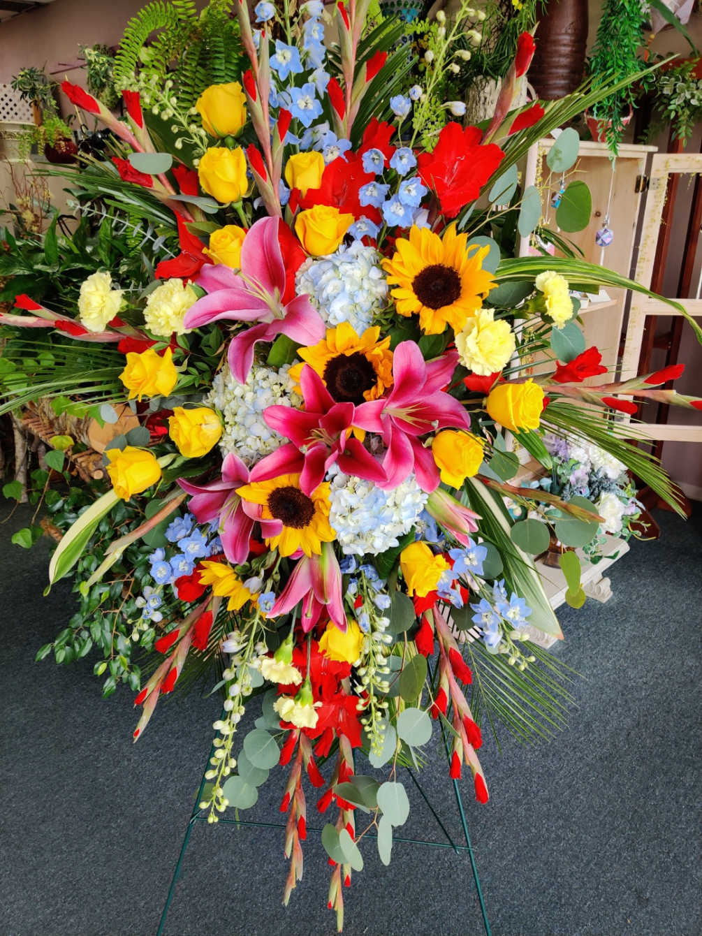 Easel spray that contains Gladiolas, Delphinium. Sunflowers, Roses, Hydrangeas, Roses, Carnations, and