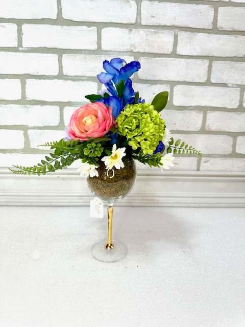 A wine glass arranged with artificial coral ranunculus, green hydrangea, lavender carnations