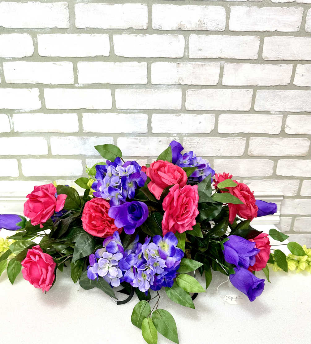 An artificial casket saddle with pink and purple roses, periwinkle hydrangeas, and