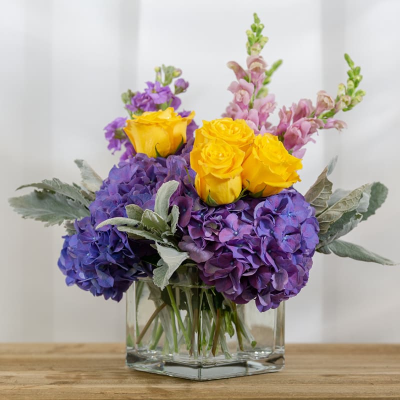 Glass cube of yellow roses, purple hydrangea, lavender snap dragon, purple stock.