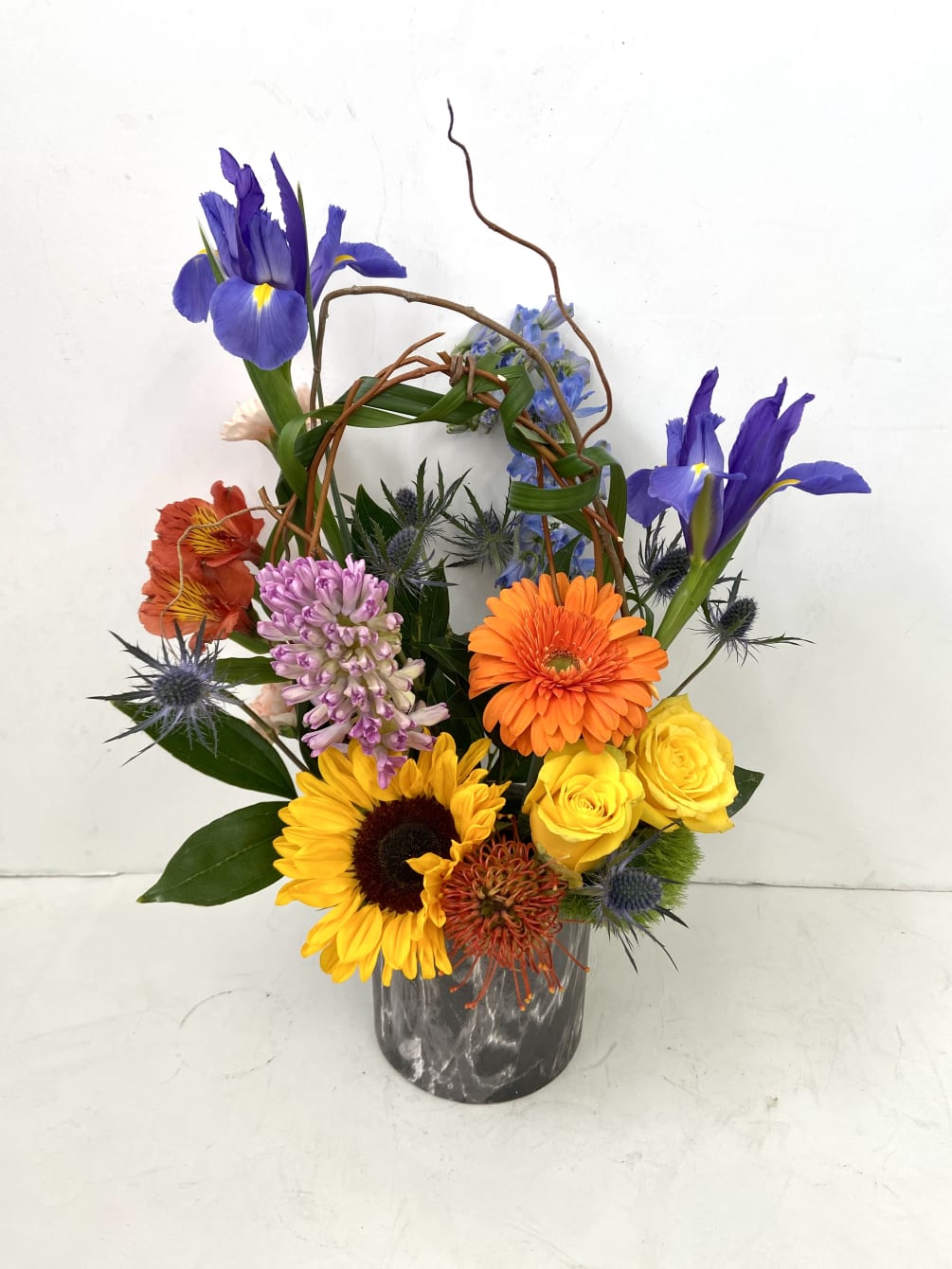 cylinder with springy mix of iris, hyacinth, roses, gerbera, sunflower, delphinium and