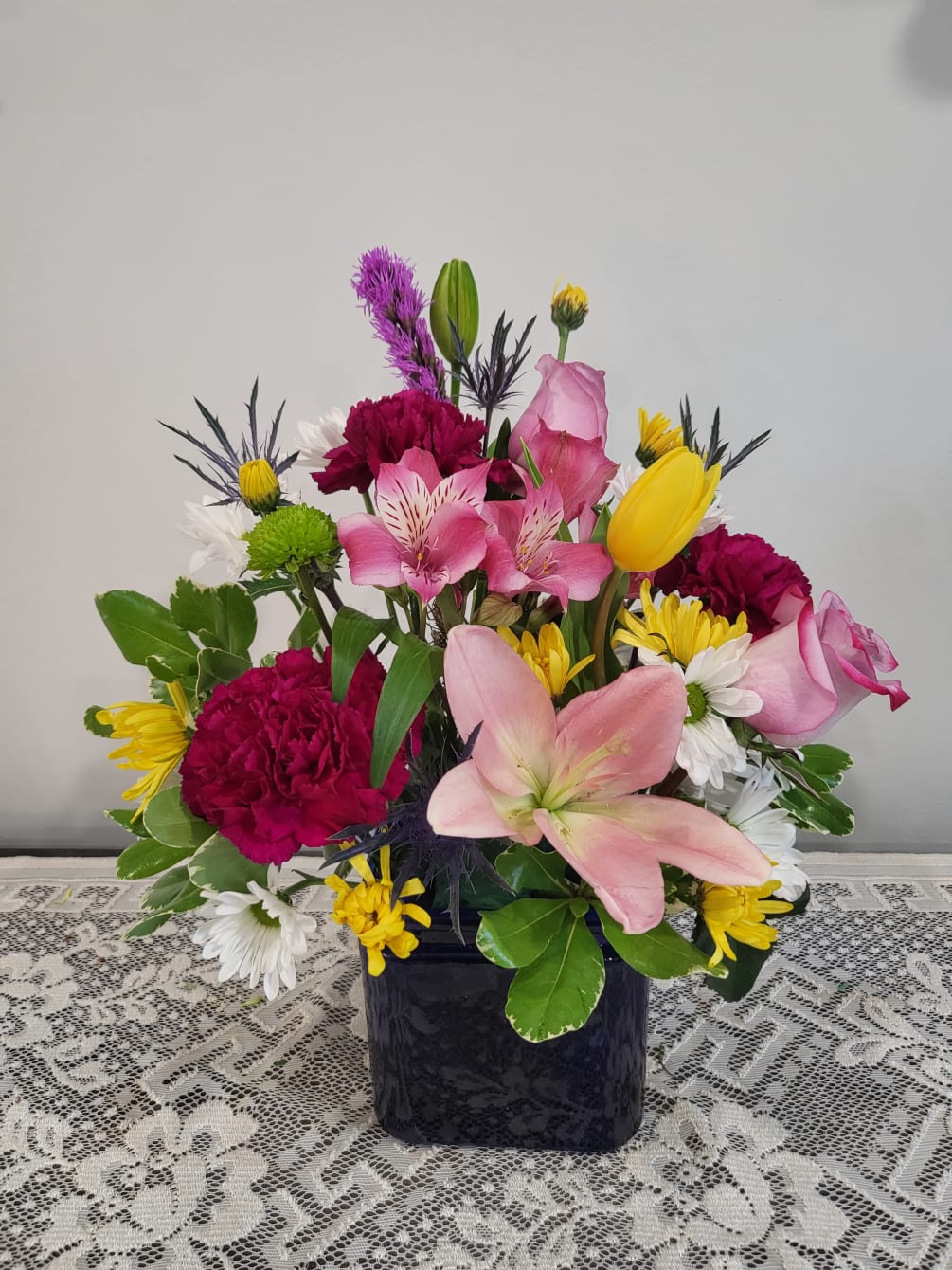 Iris,lilies,carnations,tulip and poms designed in a beautiful blue glass container with greens