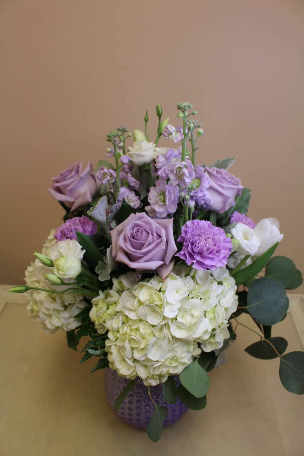 A lavender iridescent vase filled with hydrangea, purple roses and other flowers