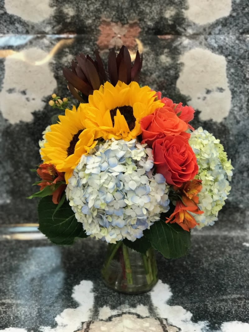This warm arrangement was designed with Sunflowers, Orange Roses, Sunset Safari Protea