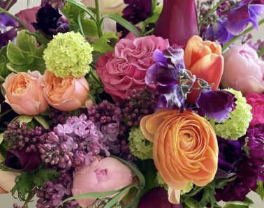 Vibrant mixed arrangement with pink roses, orange ranunculus, tulips, and purple blooms.