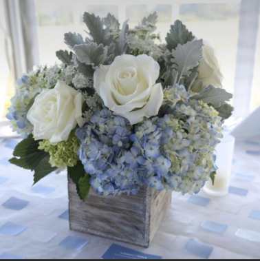 White roses and blue hydrangeas in a rustic wooden cube vase on a softly patterned table.