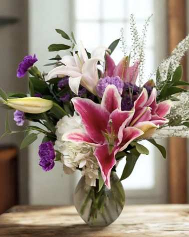 Pink lilies, white hydrangea, and purple flowers arranged in a clear glass vase on a wooden table