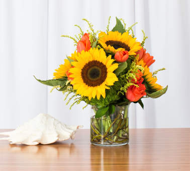 Yellow sunflowers and orange roses in a clear glass vase on a table with a white seashell.