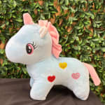 Pastel unicorn plush toy with heart patches on a leafy backdrop