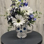 White daisies and blue flowers arranged in a skull mug with a black-and-white ribbon