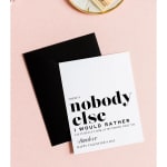 White Valentine card with bold black text and black envelope on a pink surface