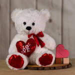 White teddy bear holding a red heart and ribbon beside a small heart decoration