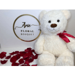 White teddy bear with a red ribbon beside rose petals and a floral bouquet sign