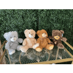 Four teddy bears sit on a glass table against a green hedge wall.