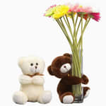 Two teddy bears beside a glass vase of colorful daisies