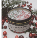A scented candle in a black tin surrounded by cranberries and pine sprigs