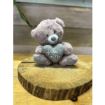 Small mauve teddy bear holding a gray heart pillow, sitting on a round wood slice.