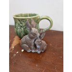 Two small rabbit figurines sit in front of a green ceramic mug.