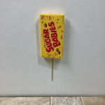 Yellow Sugar Babies candy box attached to a wooden stick against a light wall.