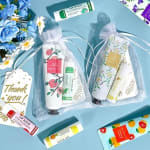 Gift bags with lip balms and hand creams on a blue surface beside blue and white daisies.