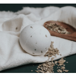 White bath bomb with herbal flecks on a towel beside a wooden scoop of dried leaves