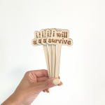 Hand holding wooden cake toppers that spell "I will survive"