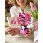 Pop-up pink and purple birthday card shaped like balloons in a gift box, held beside an open greeting card