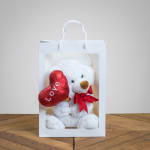 White teddy bear in a gift bag holding a red heart balloon