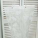 Sheer white curtain with a floral pattern hanging in front of blinds