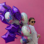Woman holding purple and birthday balloons against a pink wall